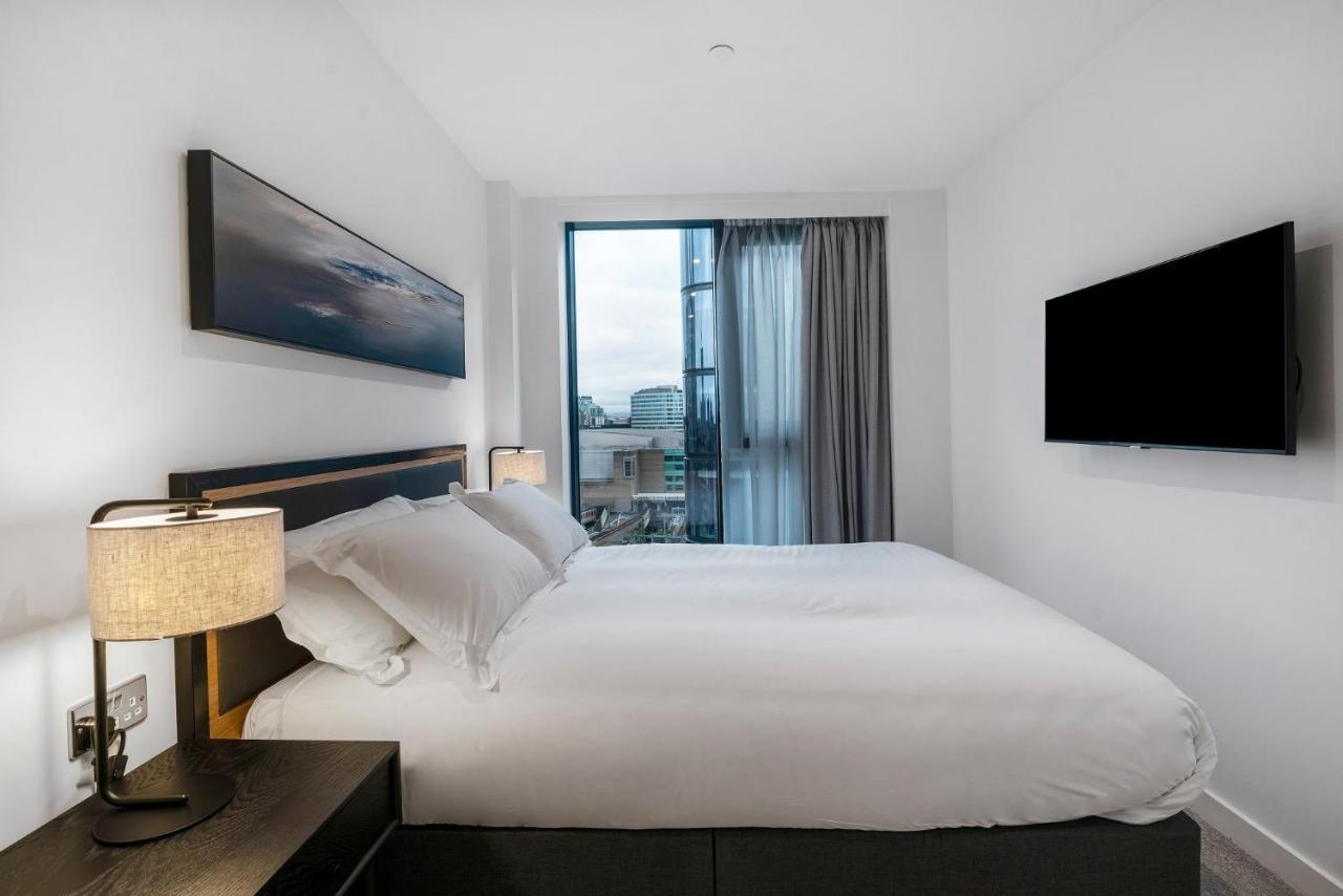 Citysuites 2 Aparthotel Mánchester Exterior foto The photo shows a modern bedroom with a minimalist aesthetic. It features a large bed with white bedding and two pillows. On either side of the bed, there are bedside tables with lamps. A large window lets in natural light, covered by sheer curtains.